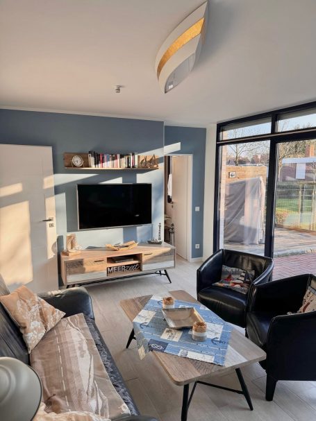 Wohnbereich Wohnzimmer mit großer Glassfront. Ein Doppelsofa und zwei Sessel, Ein Smart-TV und ein Tisch sind dort zu finden.