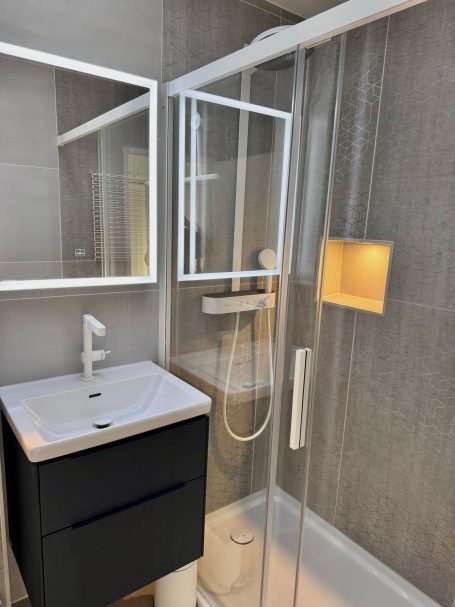 Bad Ein Badezimmer mit Waschtisch mit 2 Schubladen, Ein großer Spiegel inklusive Beleuchtung, eine Glaswand hinter der eine Regendusche ist.
